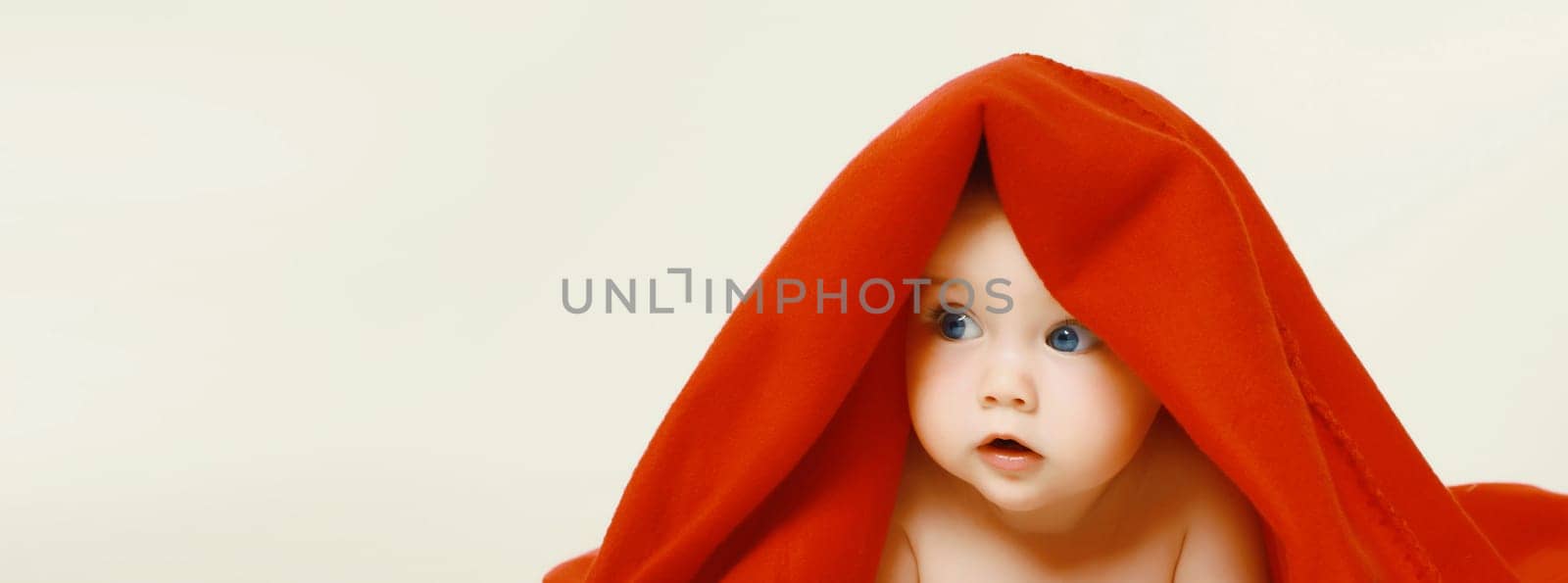 Portrait of cute baby lying under red bath towel looking away on white studio background