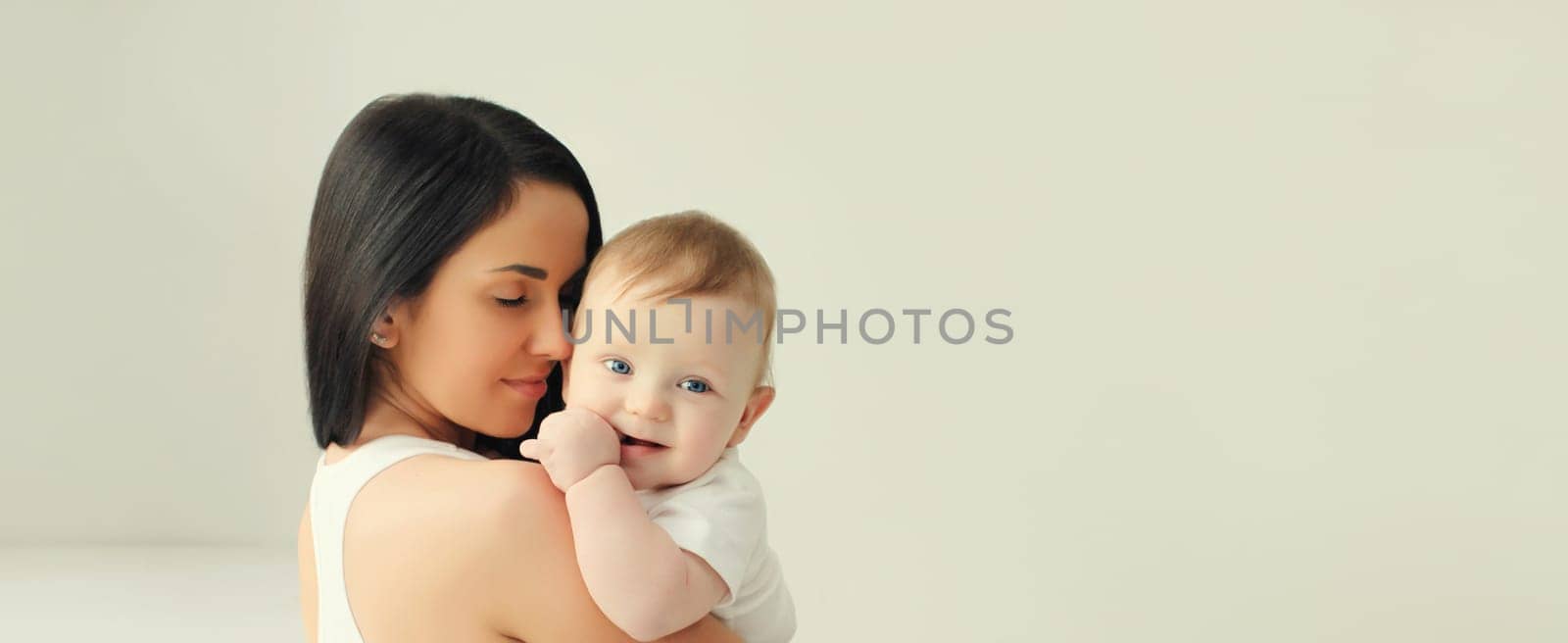 Happy young mother holding cute baby together in white room at home by Rohappy