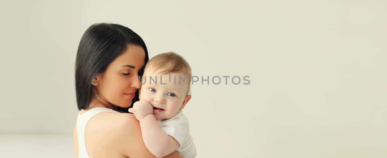 Happy young mother holding her cute baby sitting together in white room at home