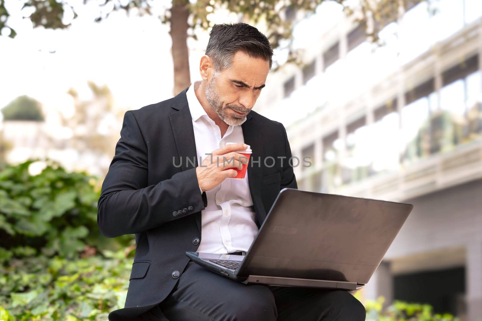 A middle-aged adult man in a suit, drinking coffee, looks at his laptop.