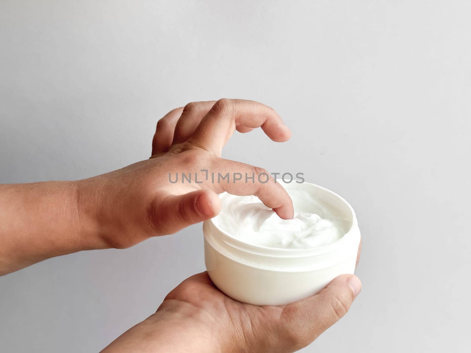 Close up of childs hands holding container of hand cream and dipping finger on white background. Skincare and moisturizing concept. Design for healthcare, wellness, and child care poster. High quality