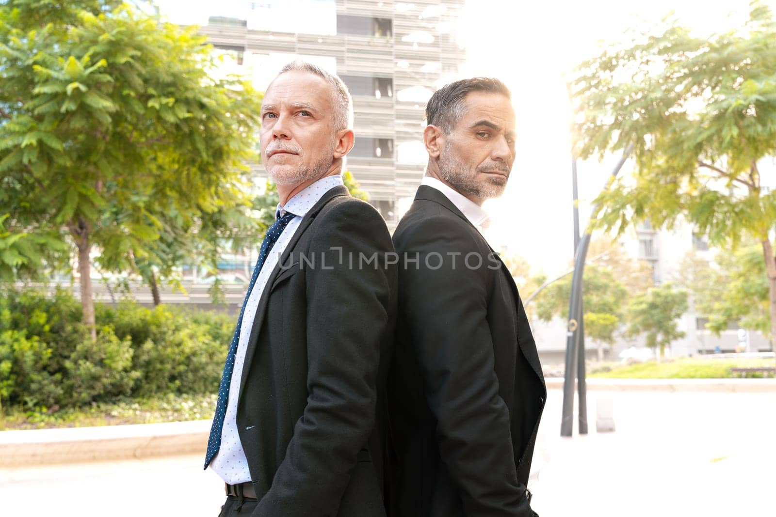 portrait two successful businessmen standing back to back looking at camera. by mariaphoto3