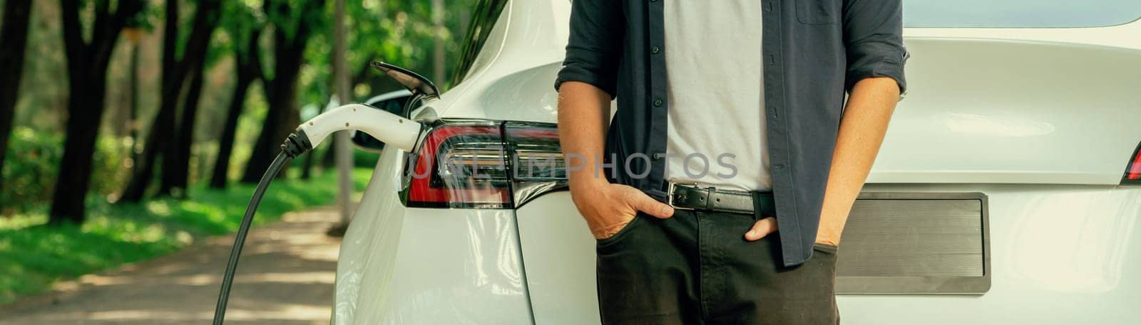 Man recharging battery for electric car during road trip travel EV car in natural forest or national park. Eco friendly travel during vacation and holiday. Panorama banner Exalt