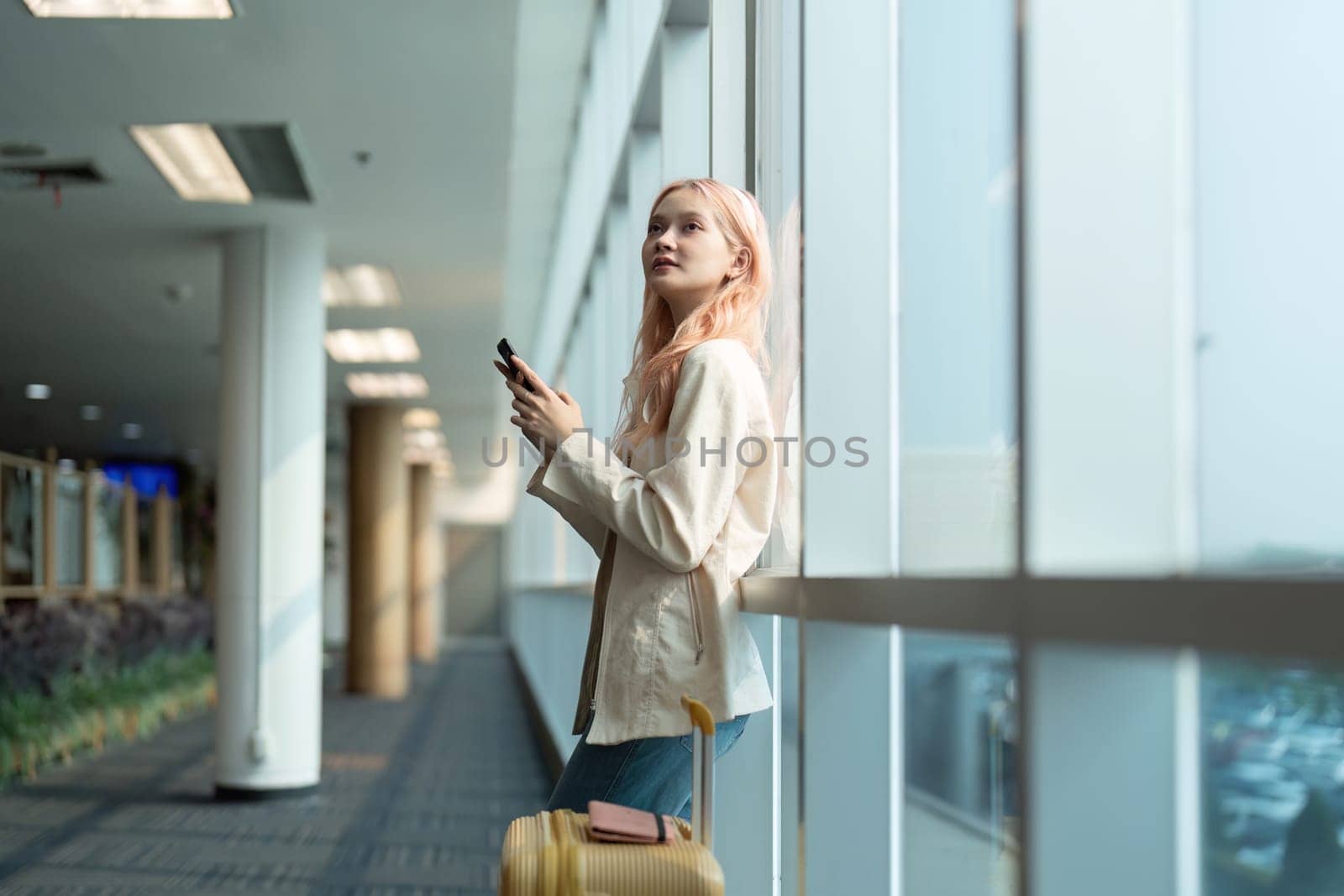 Young woman with suitcase using smartphone at airport terminal. Modern travel concept, casual outfit, waiting for flight.