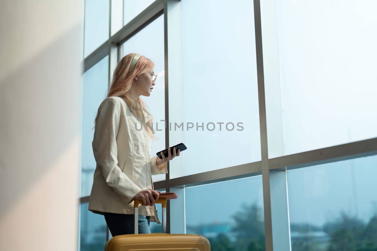 Woman traveler with luggage and phone at the airport window. Concept of travel, waiting, and departure.