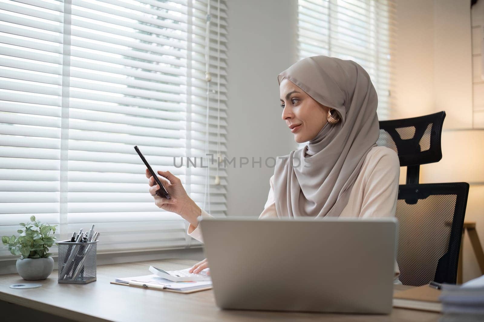 Muslim woman using smartphone at office desk. Professional workspace, casual attire.