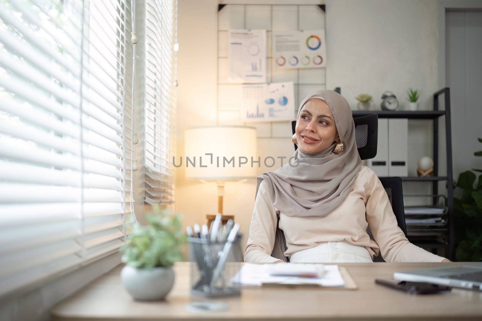 Smiling Muslim woman sitting at office desk. Professional workspace, casual attire by nateemee