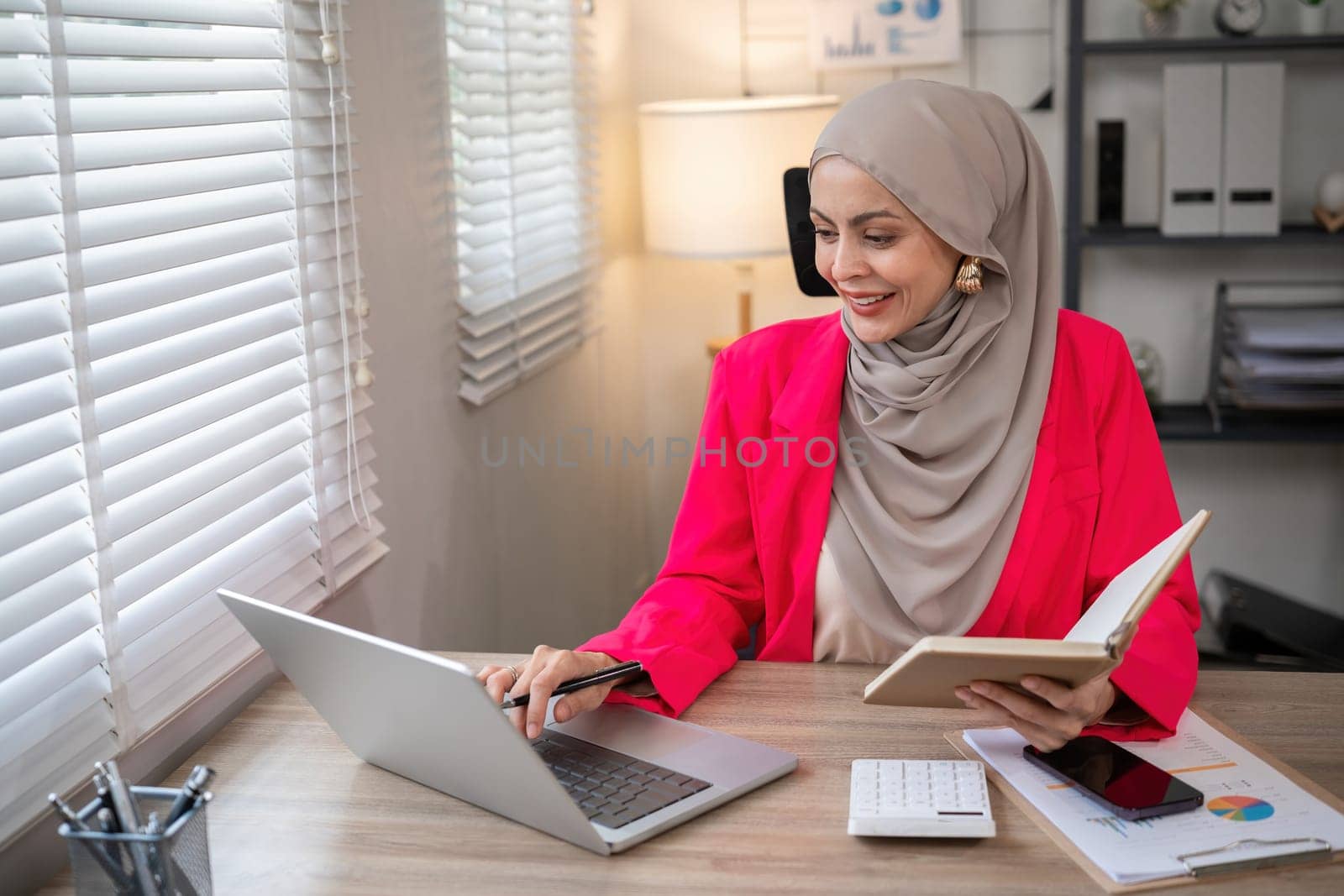 Young Muslim businesswoman wearing hijab sits working with laptop managing personal business in private office. by wichayada