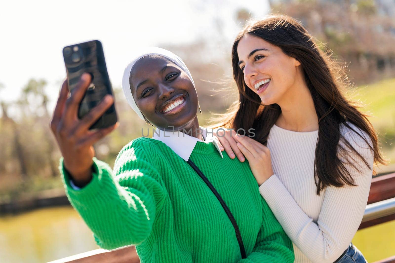 two young female friends laughing happy while having fun taking selfie photo with mobile phone in a city park, concept of friendship and modern lifestyle