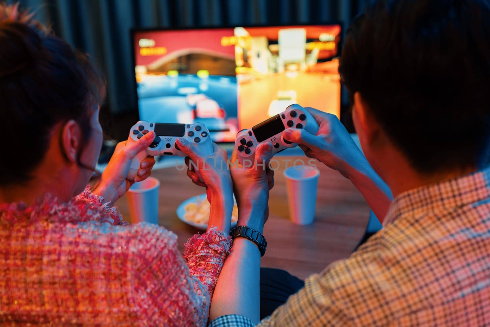 Couple gamer with controlled joystick playing car racing video game together on tv screen with snack at back side view, sitting sofa at neon light colorful living room at modern comfy home. Infobahn.