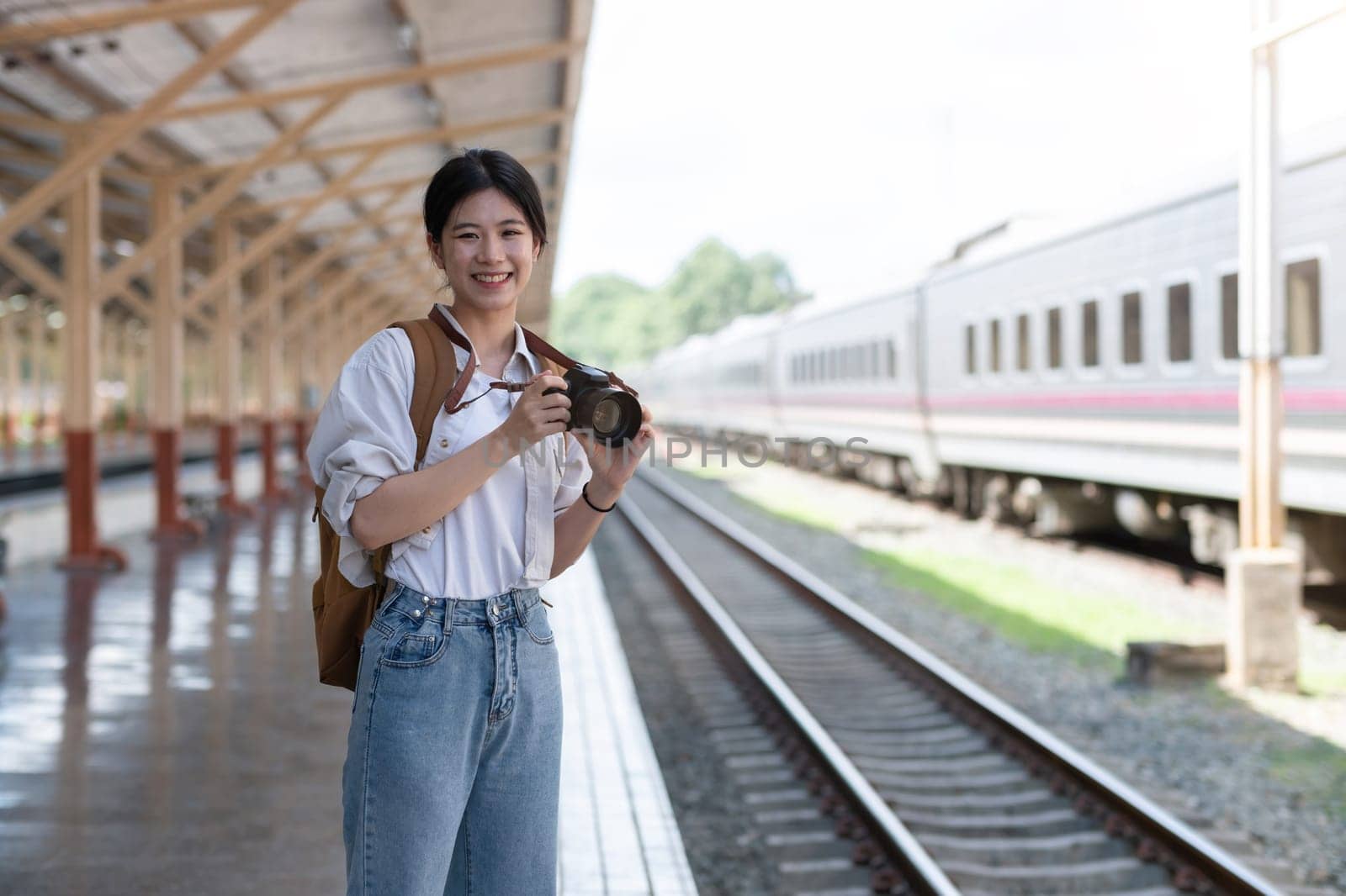 Happy young Asian female tourist carrying a backpack, holding a camera, preparing to wait for the train at the train station waiting for her vacation trip. by wichayada