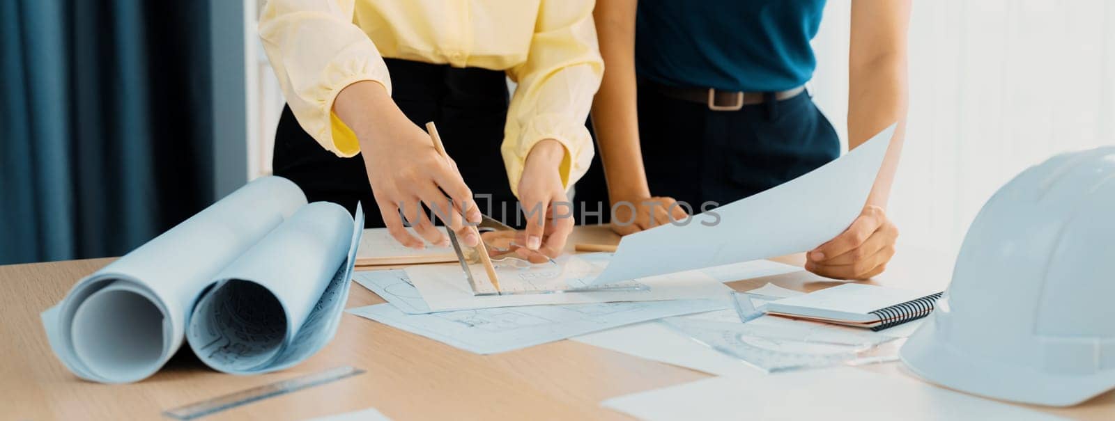 Professional architect team used divider measure during draft blueprint on table with architectural document, safety helmet and blueprint scatter around. Closeup. Focus on hand. Delineation.