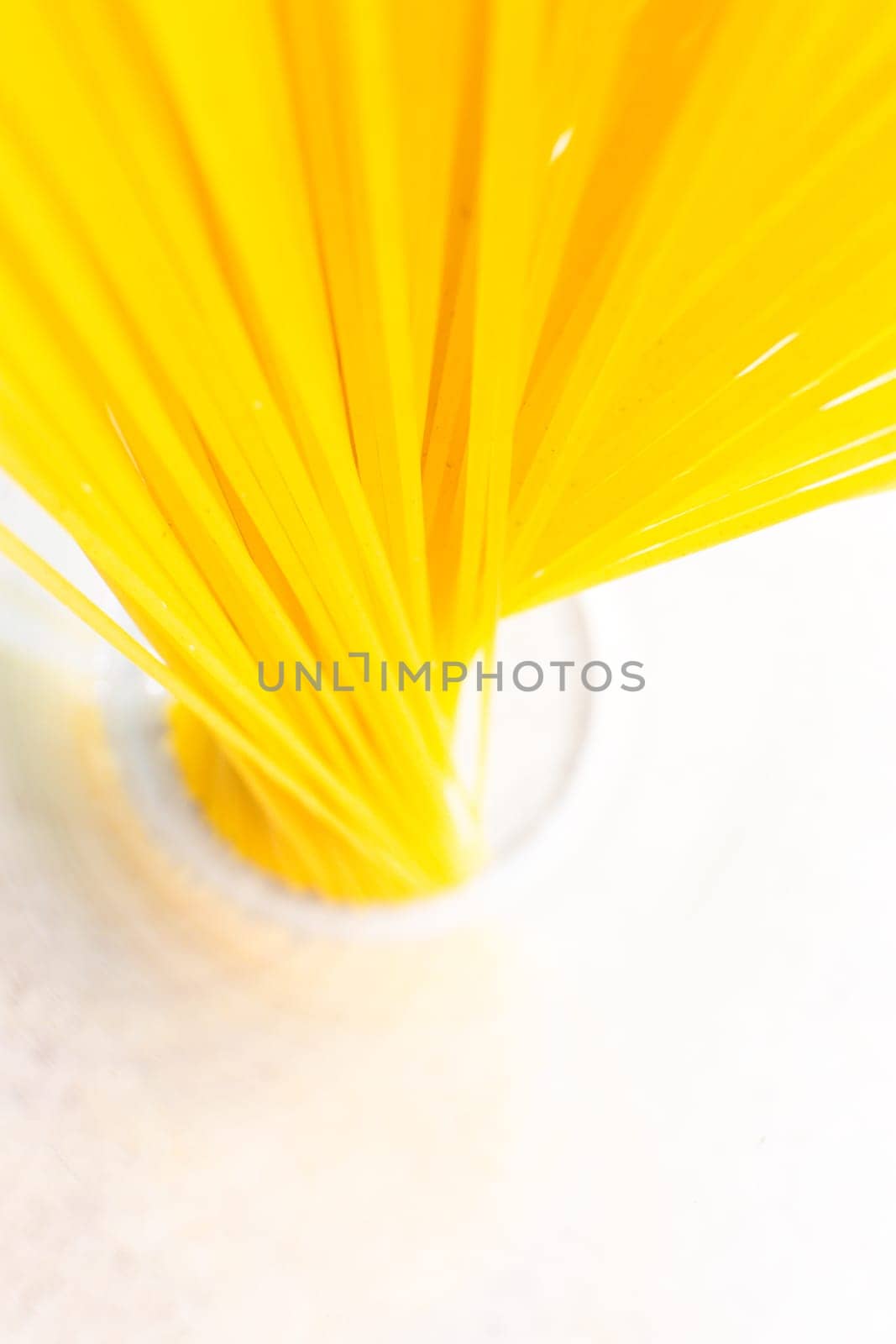 A bunch of bright yellow spaghetti arranged in a glass on a clean white surface, top view