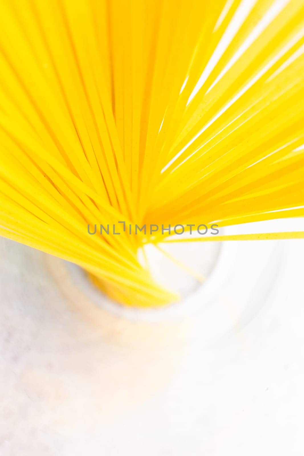 A bunch of bright yellow spaghetti arranged in a glass on a clean white surface, top view