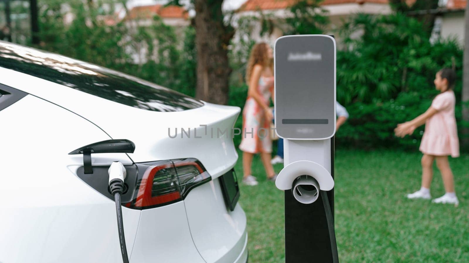 Electric vehicle recharge from home charging station on background of happy and playful family playing together. EV car using alternative and sustainable energy for better future Synchronos