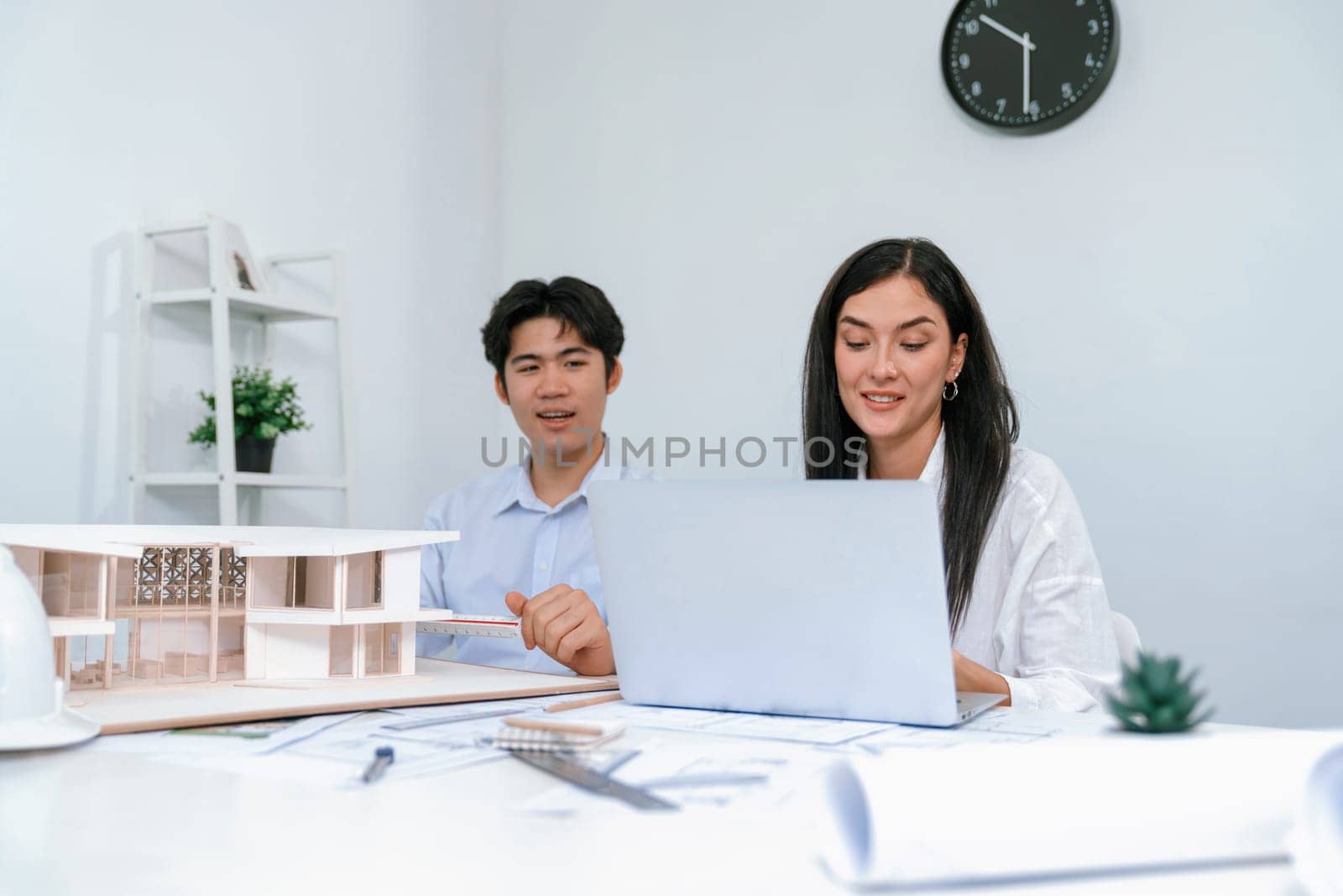 Professional male asian architect using ruler to measure house model length while young beautiful caucasian colleague using laptop to analyzed data on meeting table with house model. Immaculate.