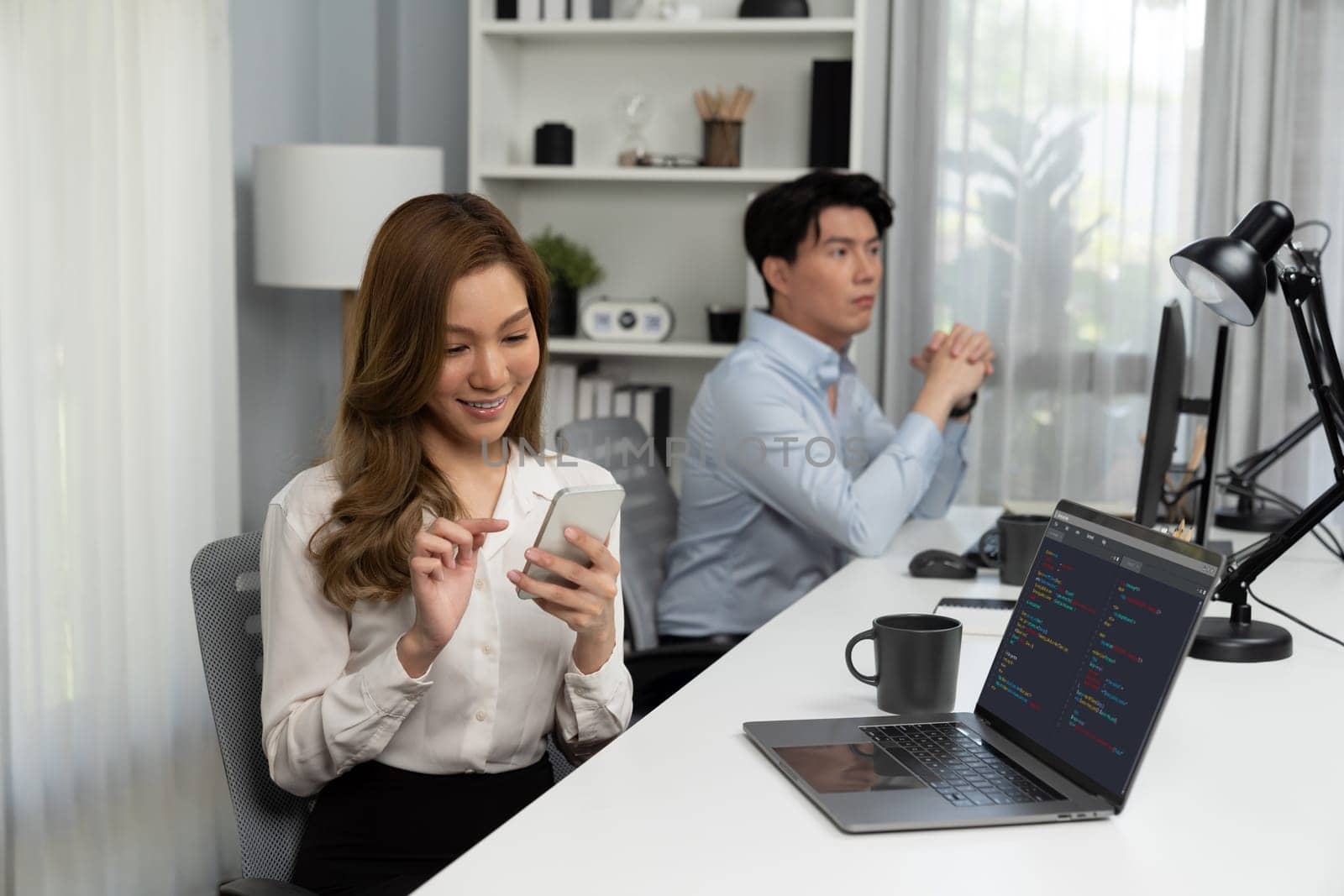 Profile smiling beautiful Asian businesswoman checking email on smartphone with laptop at modern office on working desk casual day. Blurry background man colleague analyzing data on pc. Infobahn.