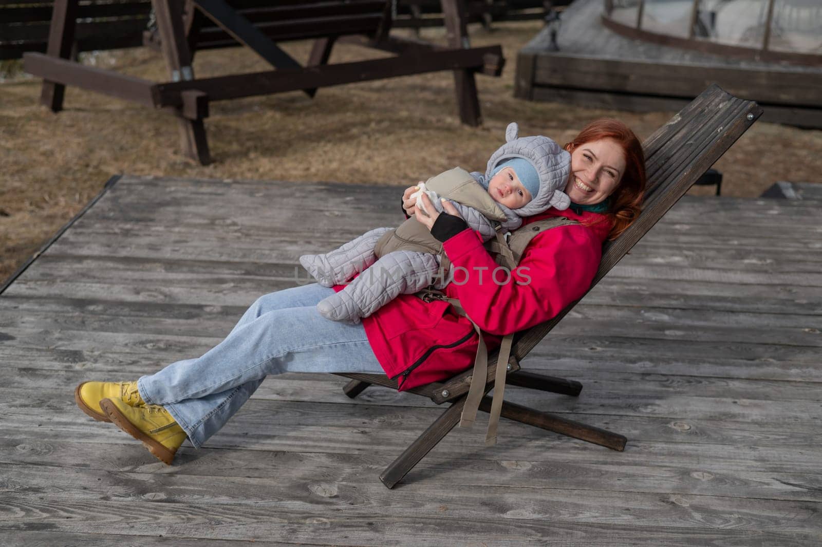 Caucasian woman with her son in an ergo backpack sitting in a wooden deck chair. by mrwed54