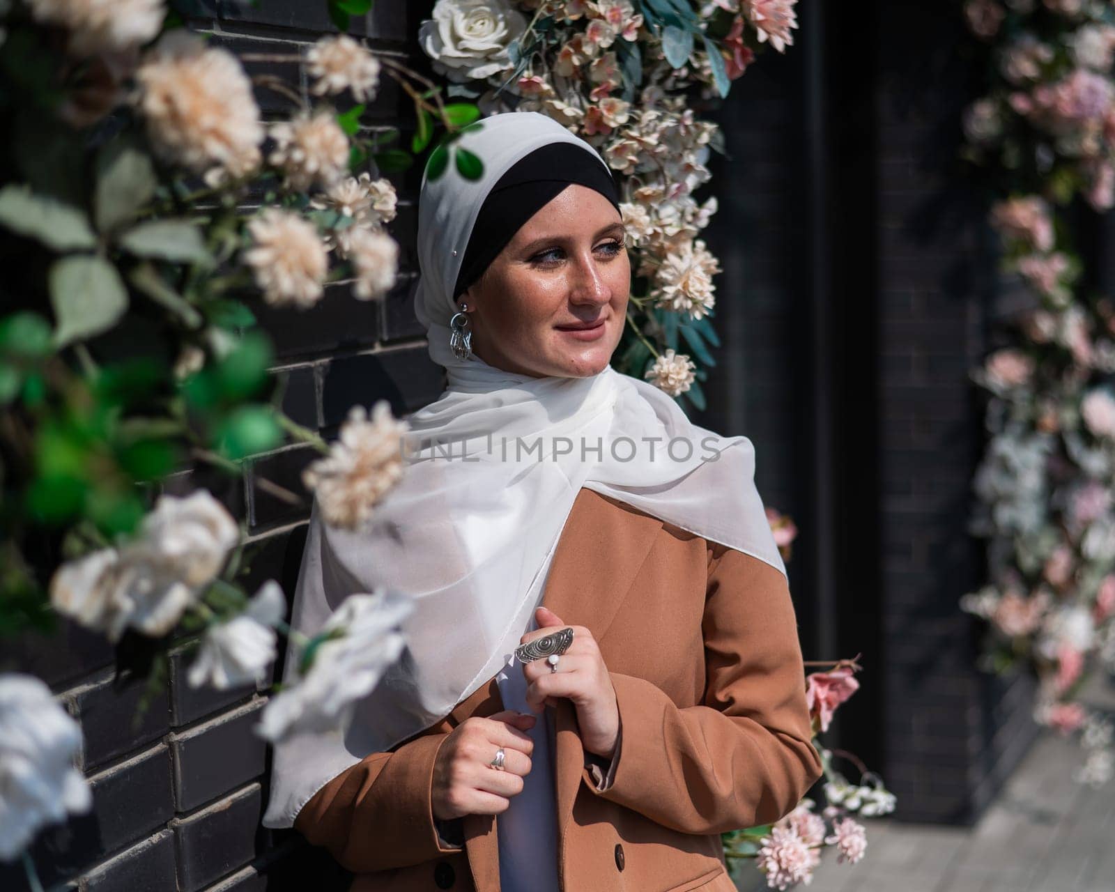 Portrait of a young caucasian woman dressed in a hijab near a wall with flowers. by mrwed54