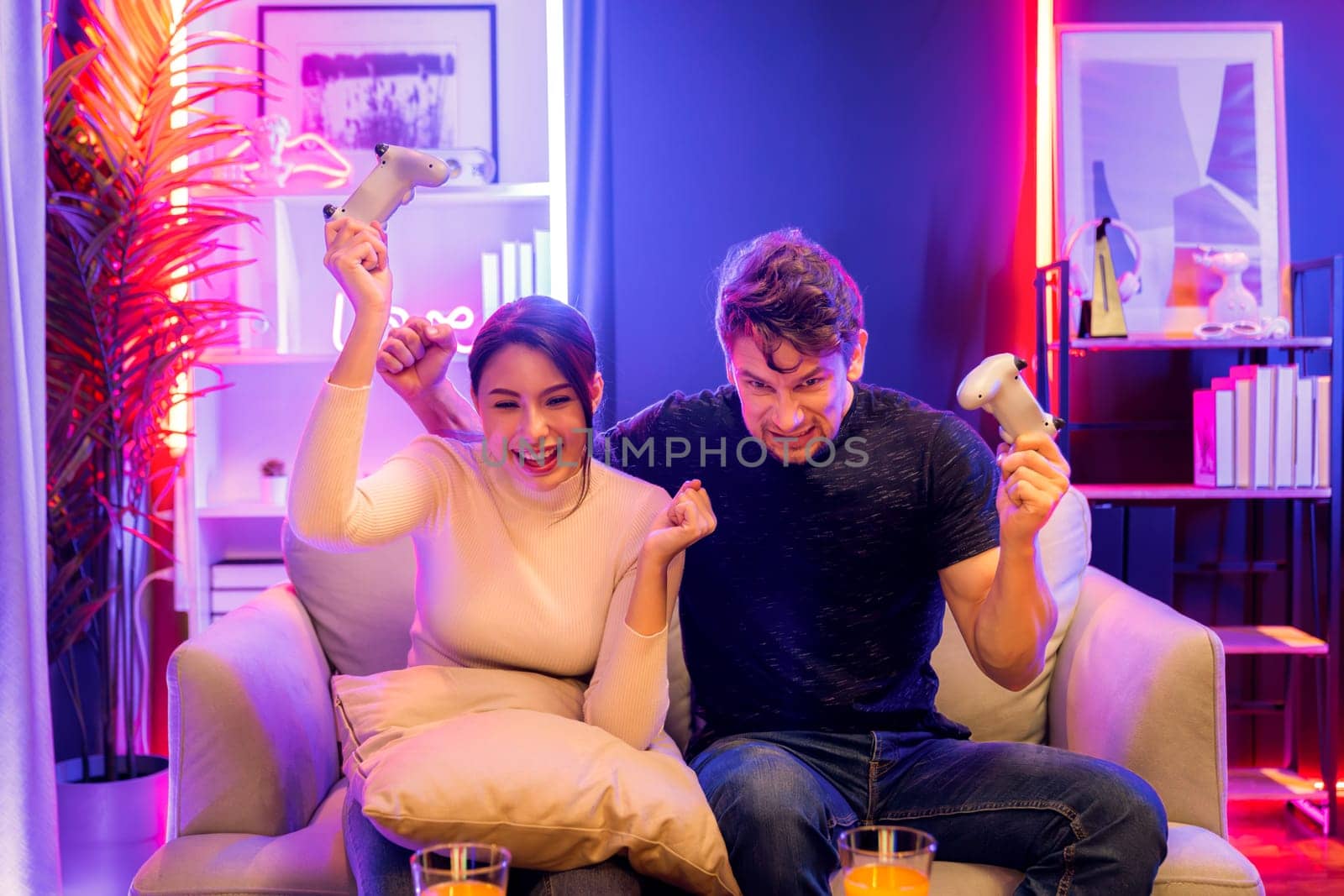 Couple joyful player video game on TV using joysticks raising fist up of winner together with snack and drinks on table in studio room in red blue neon light at comfy living home place. Postulate.