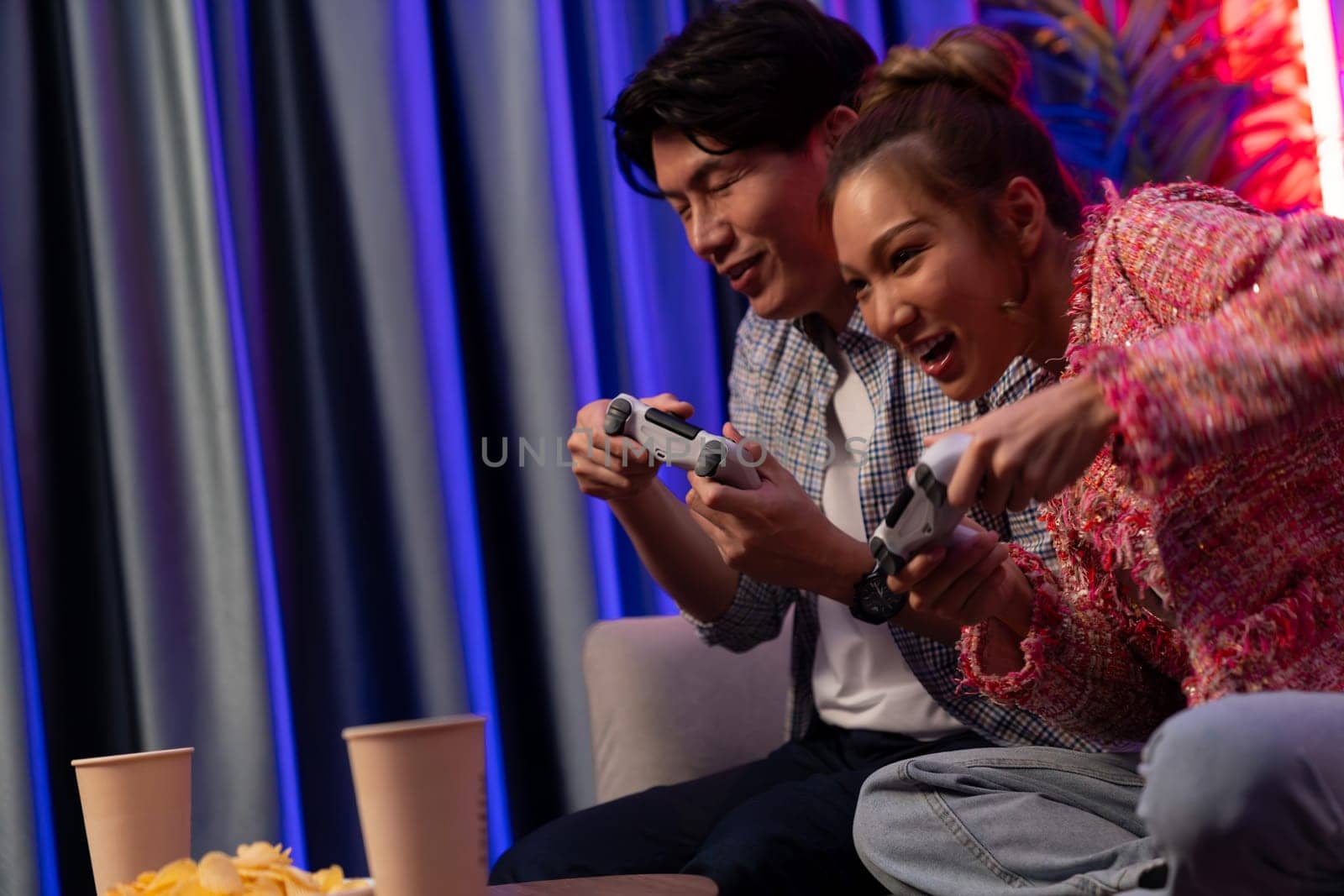 Sitting sofa of happy smiling couple playing video game control by joystick fronted snack and drinks competing challenge level with funny at modern neon blub light comfy living room at home. Infobahn.