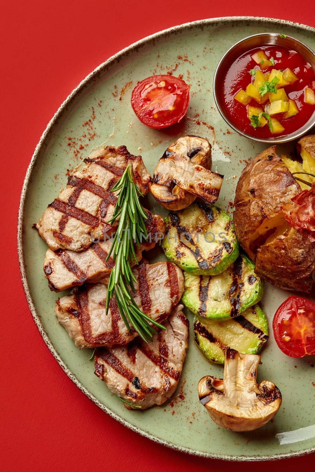 Juicy grilled pork steak seasoned with fresh rosemary served with side of with roasted veggies, jacket potato with bacon, and tomato sauce with bell pepper and greens presented on vivid red background