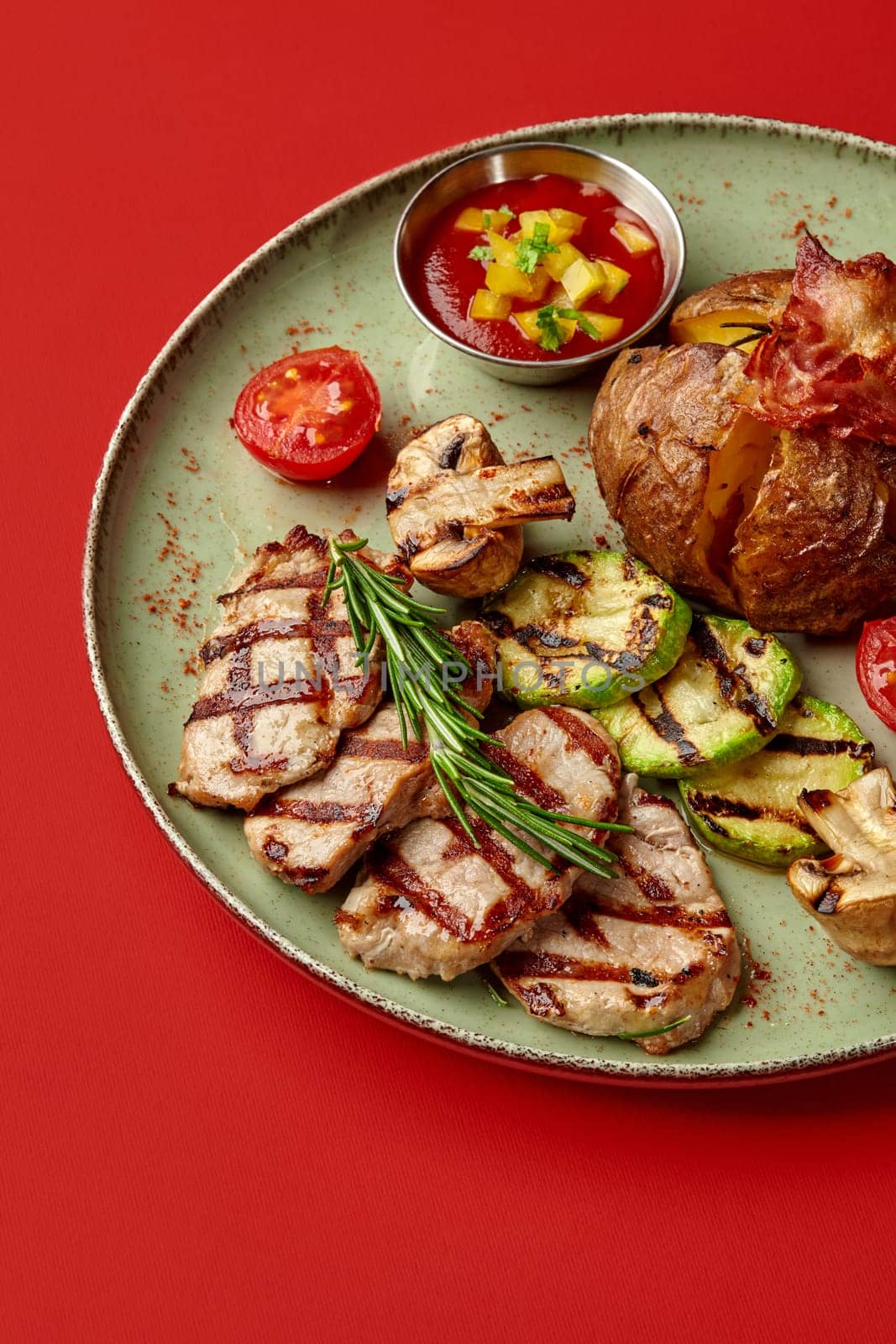 Tender grilled pork steaks with roasted zucchini and mushrooms, jacket potato topped with bacon, accompanied with tomato and yellow pepper salsa on striking red background