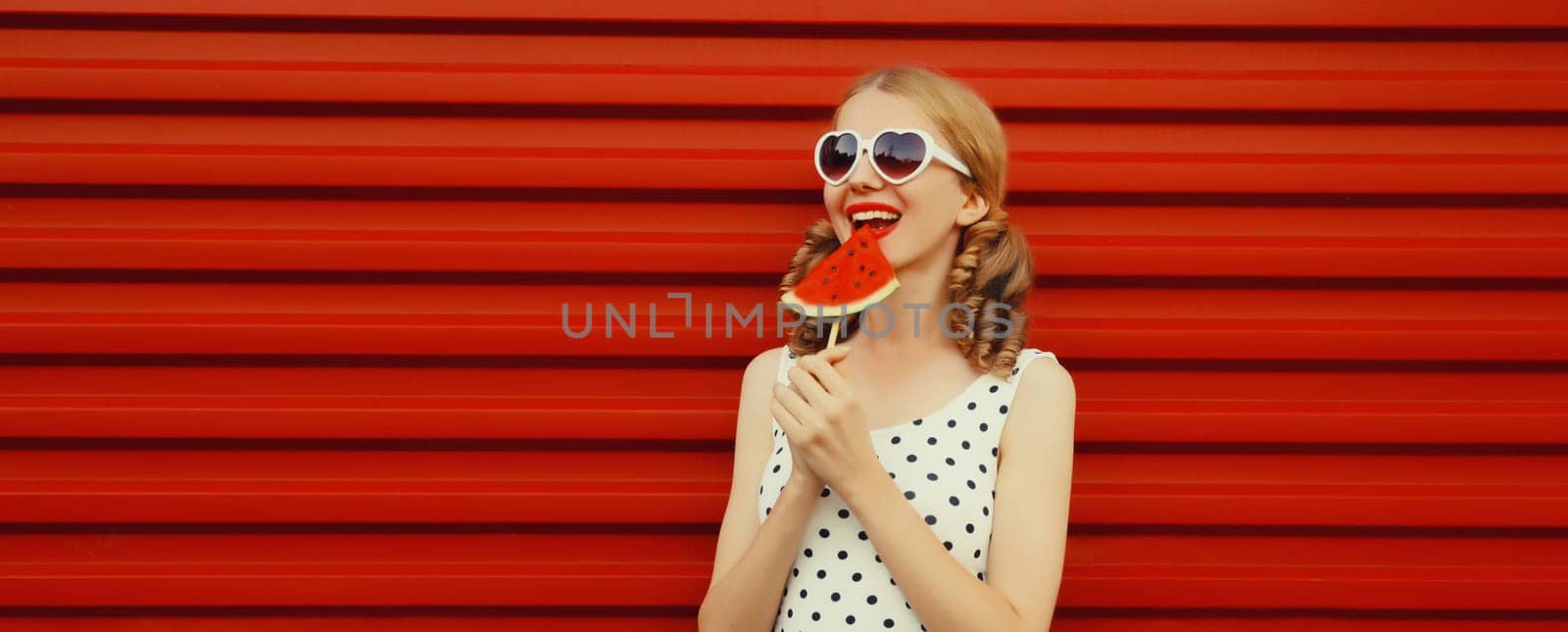 Summer portrait of happy smiling young woman with juicy fruits, lollipop slice of watermelon wearing heart shaped sunglasses with cool girly hairstyle on red background