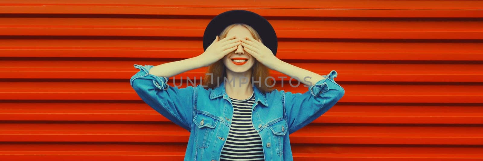 Portrait of happy surprised smiling young woman covering her eyes wearing black round hat, denim jacket on red background