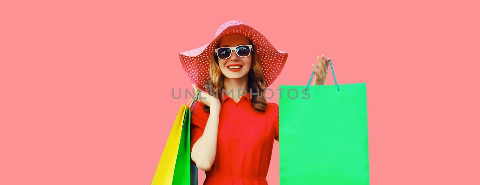 Portrait of beautiful happy smiling young woman model posing with colorful shopping bags wearing summer straw hat, dress on pink studio background
