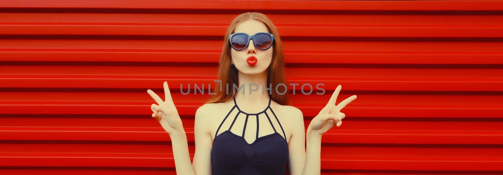 Portrait of stylish caucasian young woman model blowing a kiss wearing in black outfit, sunglasses on red background