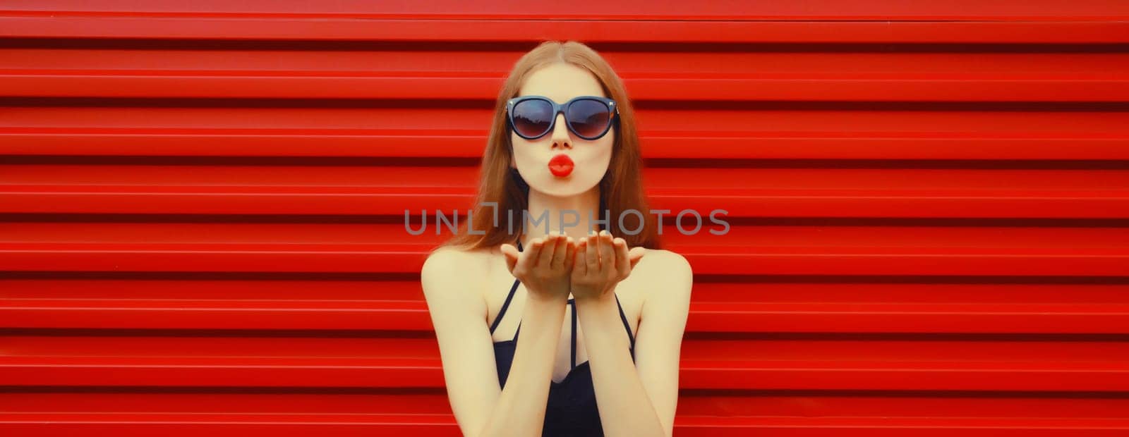 Portrait of stylish caucasian young woman model blowing a kiss wearing in black outfit, sunglasses on red background