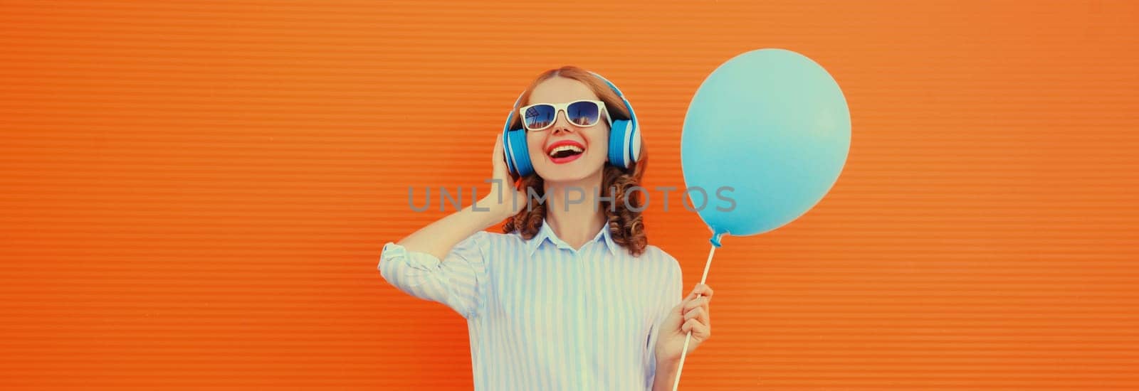 Portrait of happy cheerful young woman in wireless headphones listening to music with blue balloon on orange background