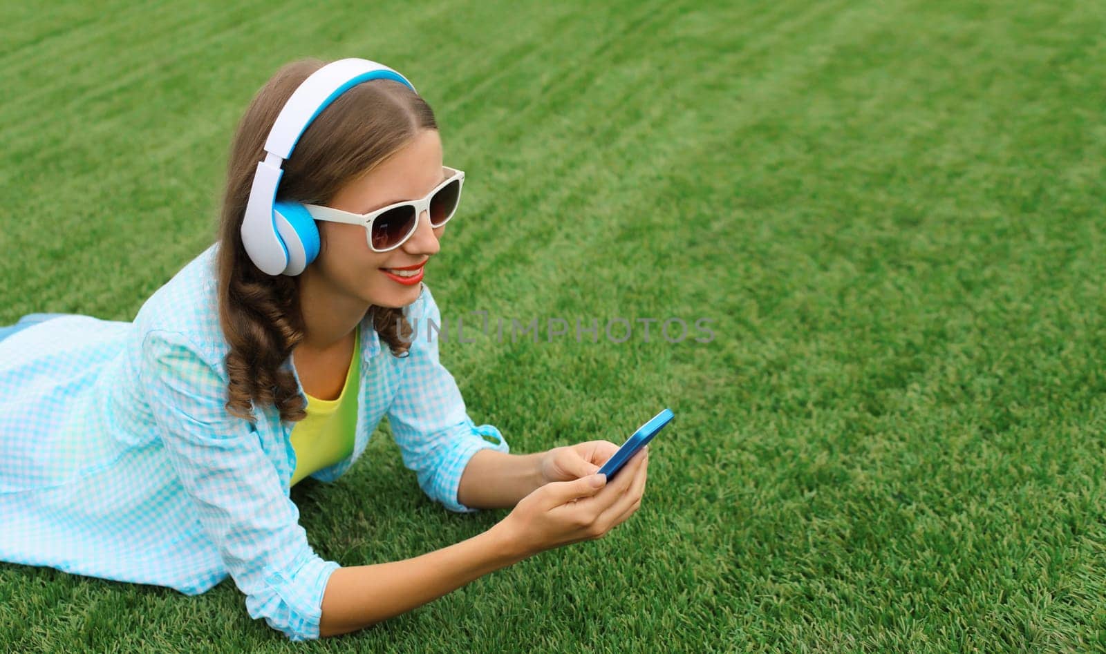 Happy young woman holding phone listening to music in headphones lying on grass in summer park by Rohappy