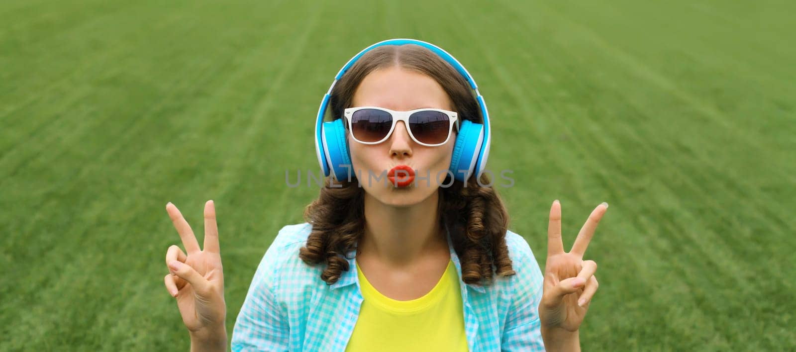 Portrait of happy smiling young woman listening to music in headphones blowing kiss while sitting on grass in summer park