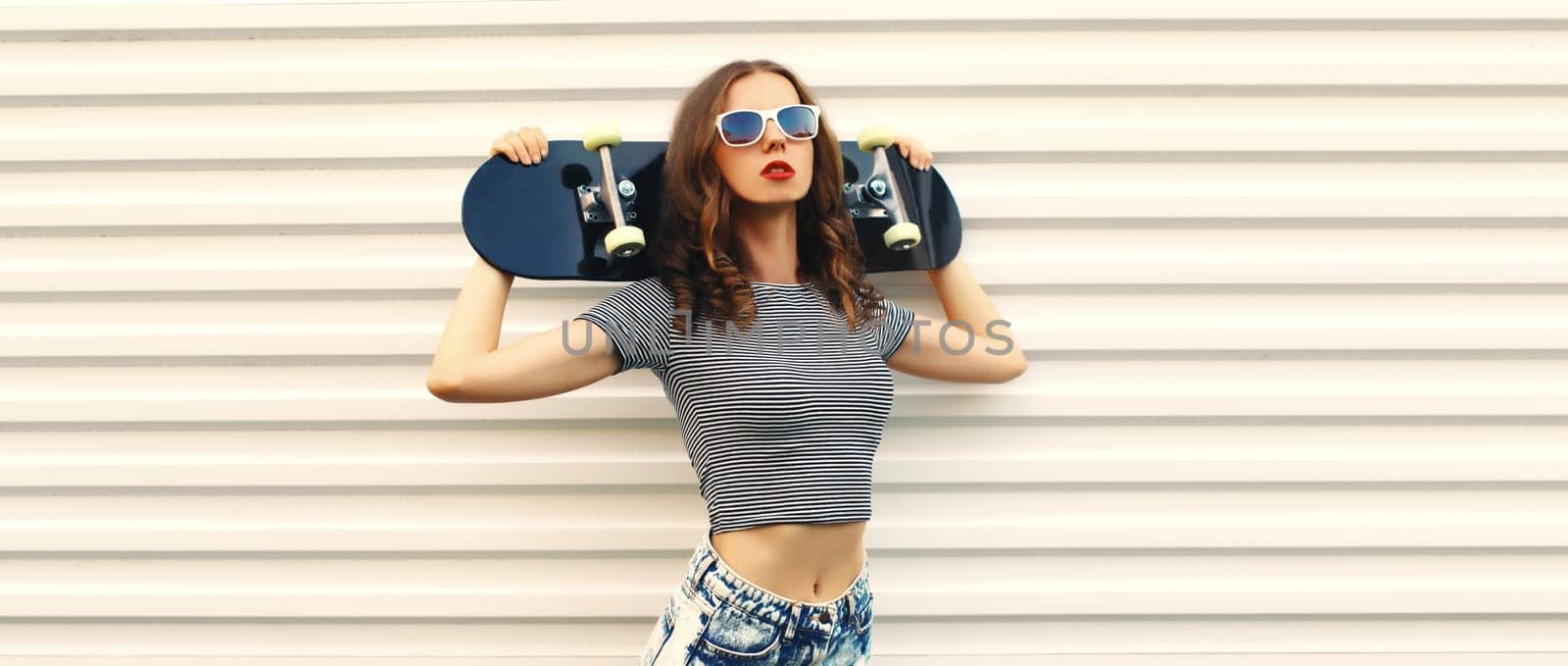 Portrait of stylish young woman posing with black skateboard on white background
