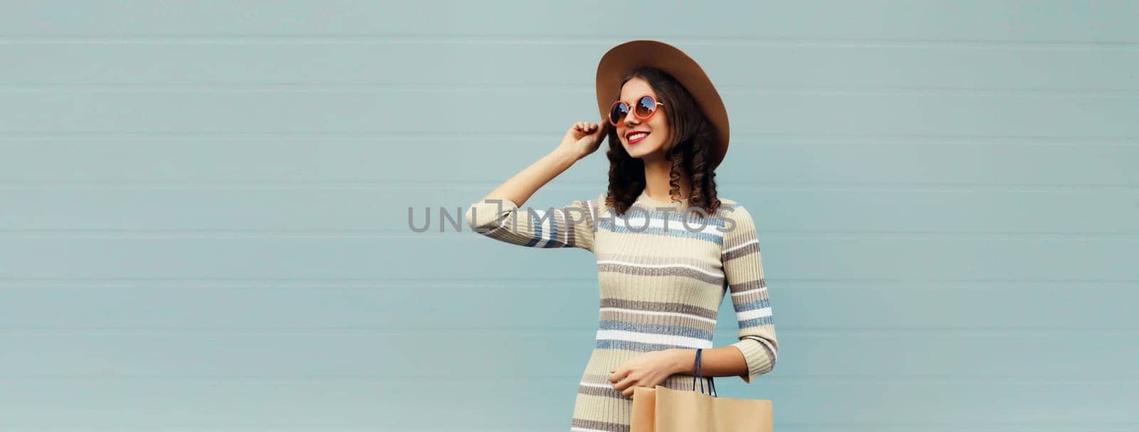 Stylish beautiful happy smiling young woman posing with shopping bags in round hat, dress on city street, gray wall background