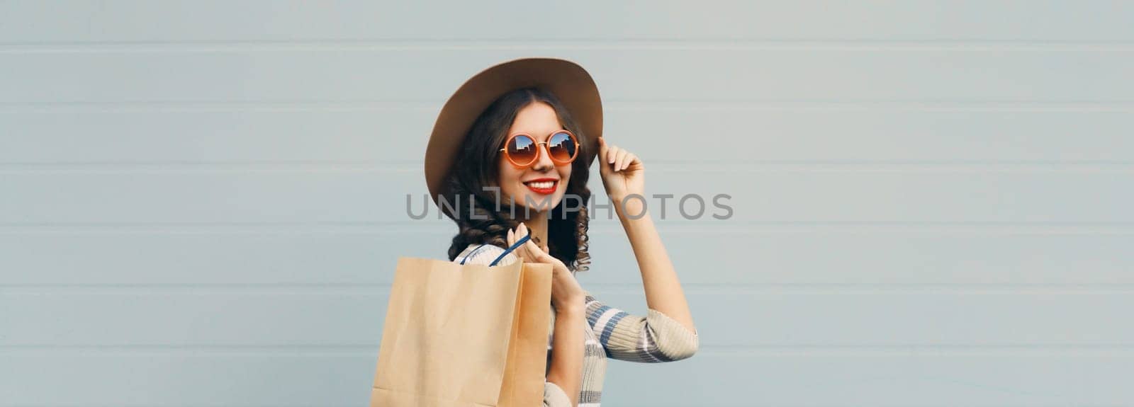 Stylish beautiful happy smiling young woman posing with shopping bags in round hat, dress on city street, gray wall background