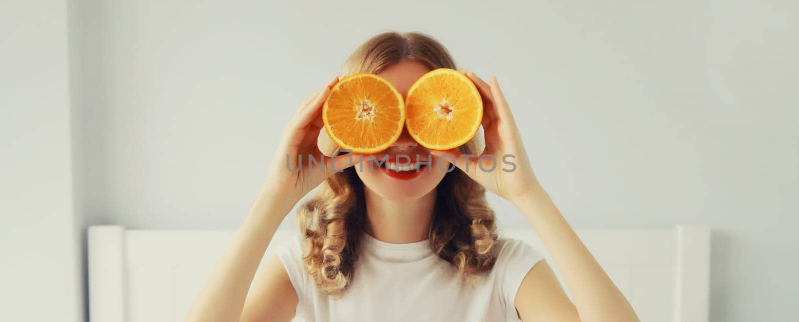 Summer, nutrition, diet and vegetarian concept. Happy healthy cheerful young woman covering her eyes with slices of orange fruits and looking for something in white room at home