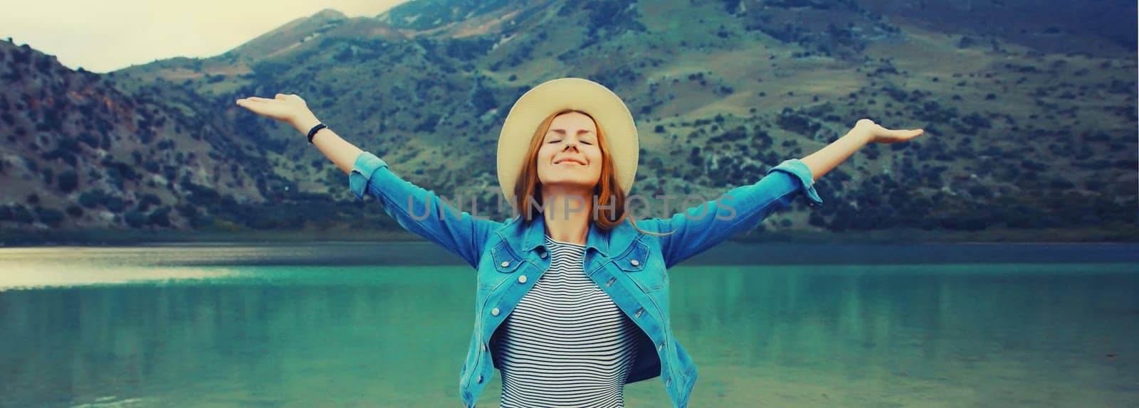 Summer vacation and tourism, rear view of happy woman tourist in straw hat raising her hands up on lake and mountains background. Greece, island Crete.