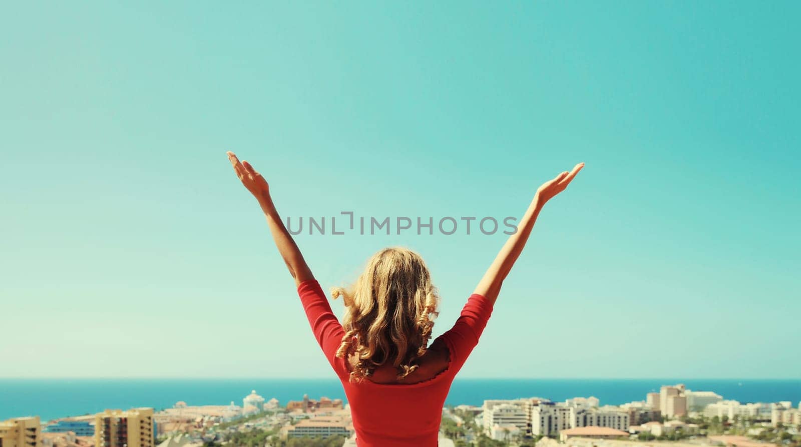 Summer vacation, beautiful happy young woman tourist raising her hands up on top of the mountain looking around the city in Tenerife, Canary Islands, Spain. Aerial top view.