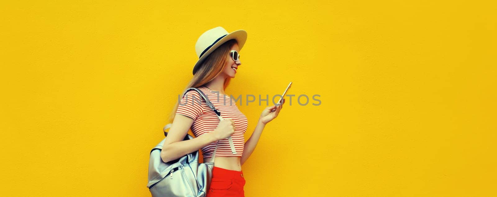 Summer portrait of happy traveler young woman 20s with mobile phone looking at device in casual straw hat with backpack on bright yellow studio background