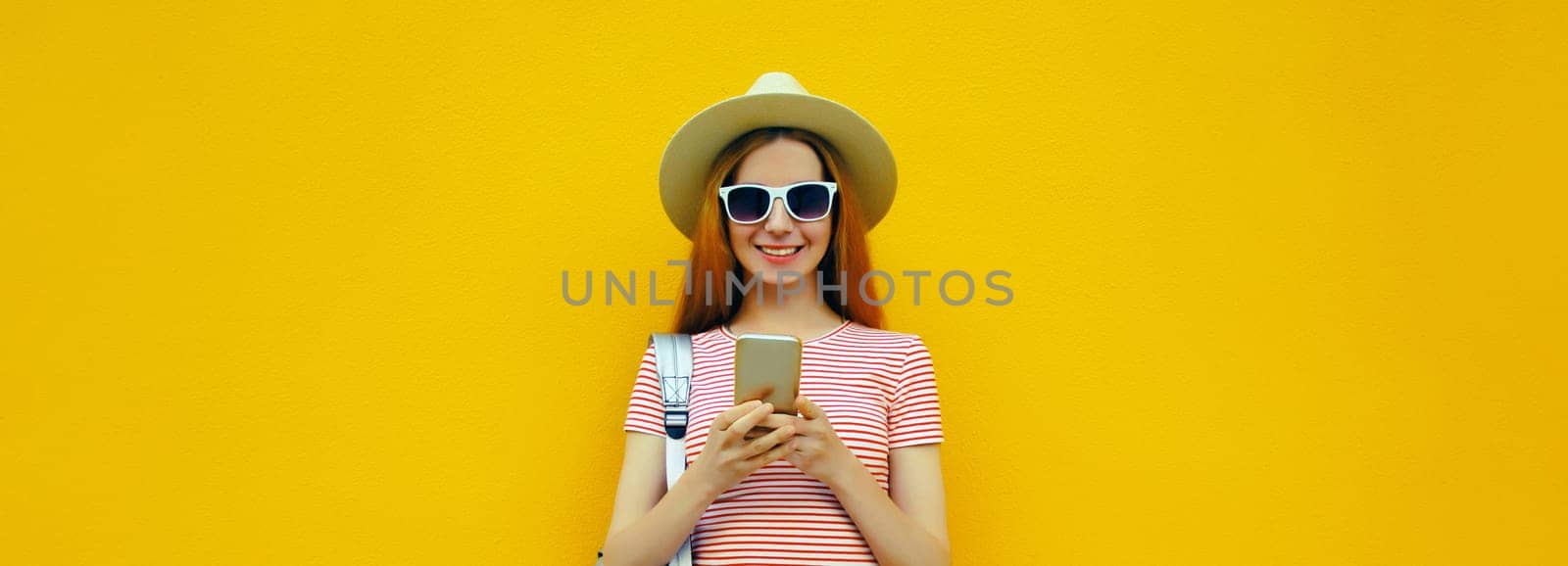 Summer portrait of happy traveler young woman 20s with mobile phone looking at device in casual straw hat with backpack on bright yellow studio background