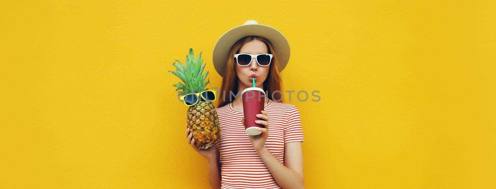 Summer bright portrait of stylish young woman drinking fresh juice holding pineapple fruit in straw hat, sunglasses on colorful yellow studio background