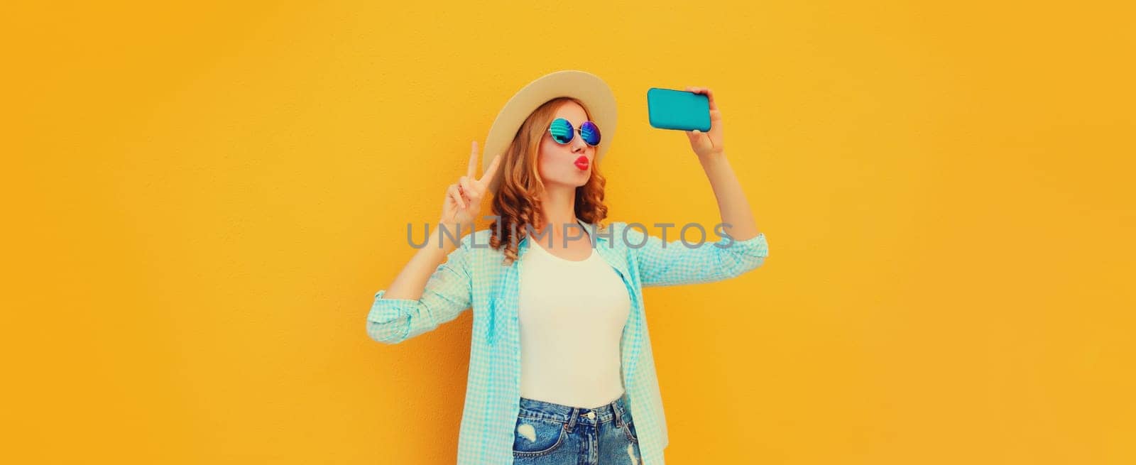 Summer cheerful stylish young woman taking selfie with smartphone blowing a kiss wearing straw hat on yellow background