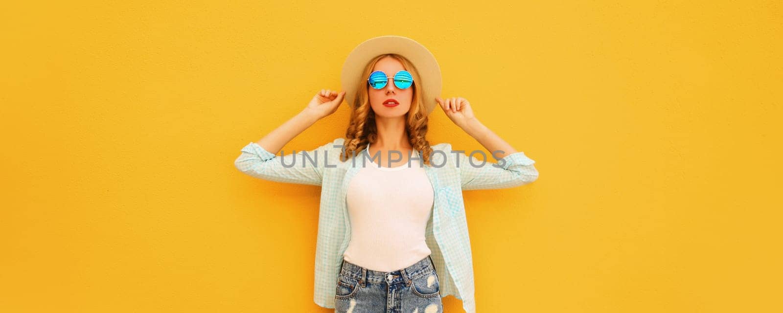 Summer portrait of stylish blonde young woman posing wearing straw hat, shirt on bright yellow studio background