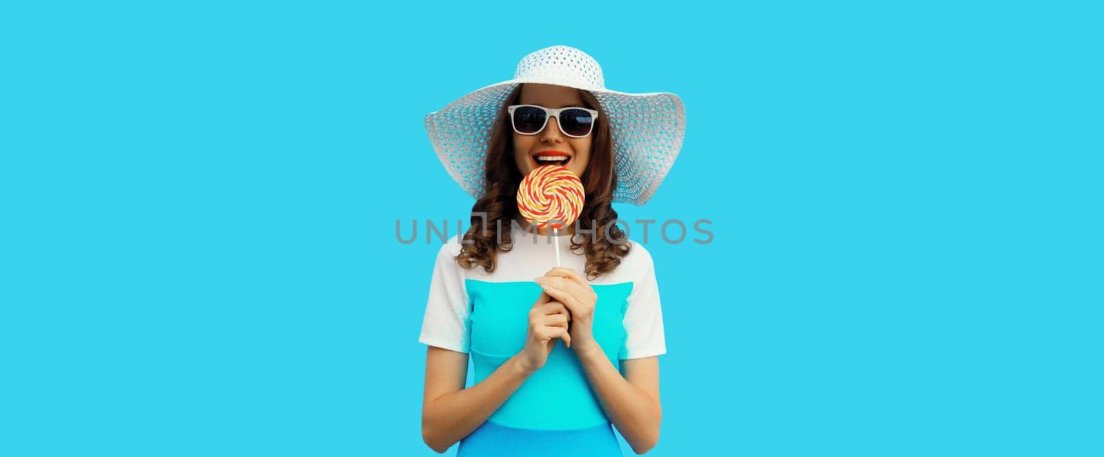 Portrait of happy cheerful smiling young woman holding colorful lollipop wearing white summer hat, sunglasses on blue background