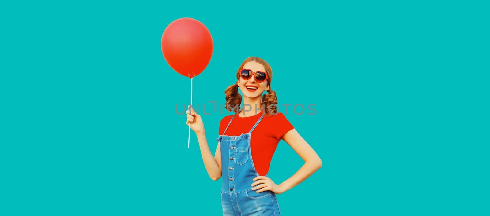 Happy cheerful young woman having fun with balloon wearing red heart shaped sunglasses, jumpsuit on blue background