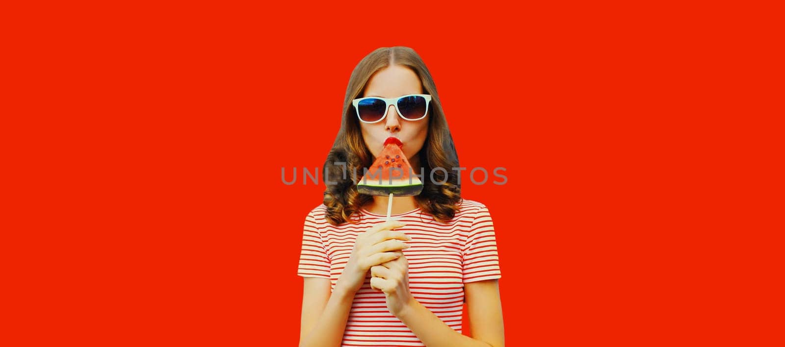 Summer portrait of stylish young woman with sweet juicy lollipop or ice cream shaped slice of watermelon wearing glasses on red studio background