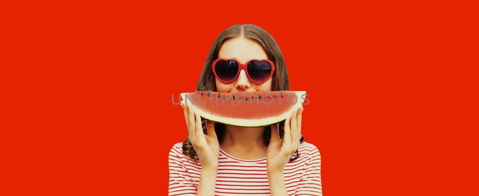 Summer portrait of happy young woman with slice of watermelon wearing red heart shaped sunglasses on studio background